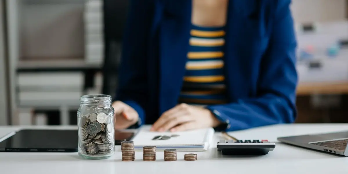 Pessoa em frente a uma mesa fazendo anotações em um caderno com algumas pilhas de moedas à sua frente