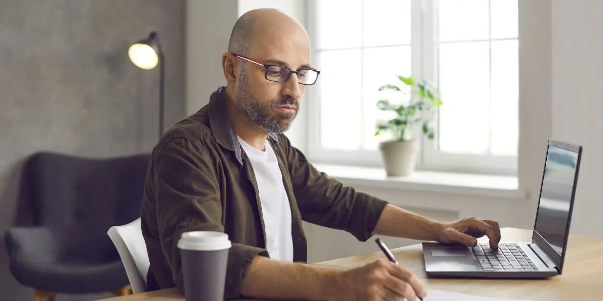 Homem sentado em frente a uma mesa usando um notebook e fazendo anotações