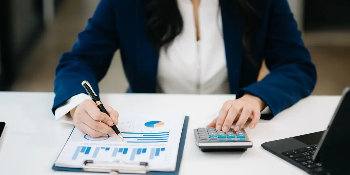 Pessoa sentada a frente de uma mesa escrevendo em papéis e fazendo cálculos com uma calculadora