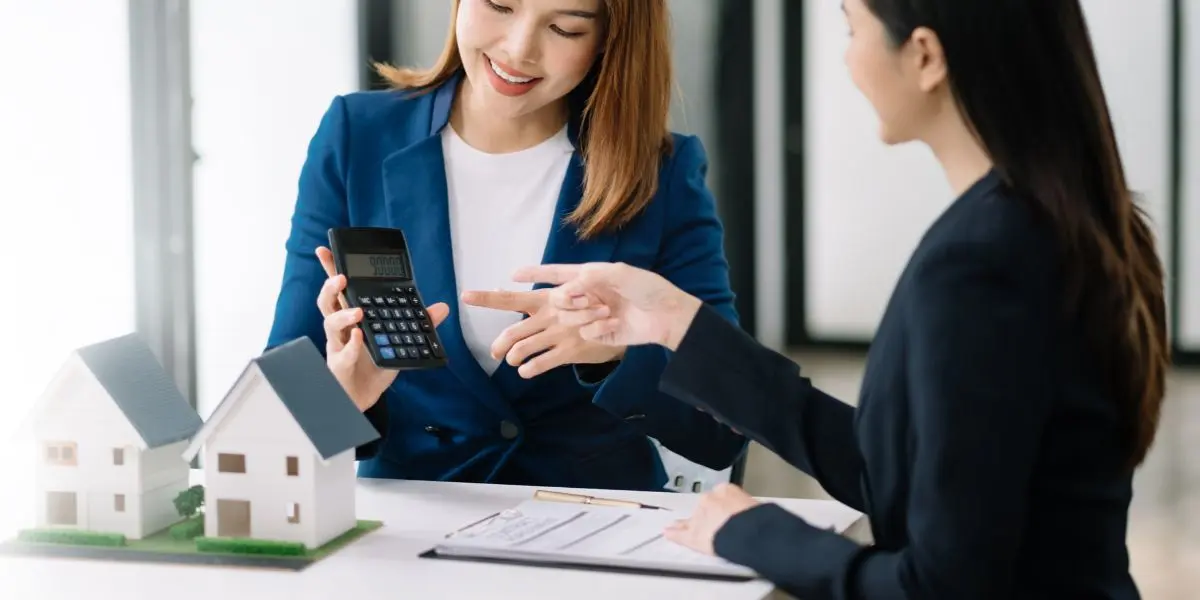 Duas pessoas olhando para uma calculadora em frente a uma mesa com a maquete de uma casa sobre ela